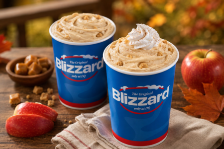 Two Dairy Queen Caramel Apple Cheesecake Blizzard treats displayed on a rustic wooden table with fresh apples, caramel pieces, and autumn leaves against a warm fall background.