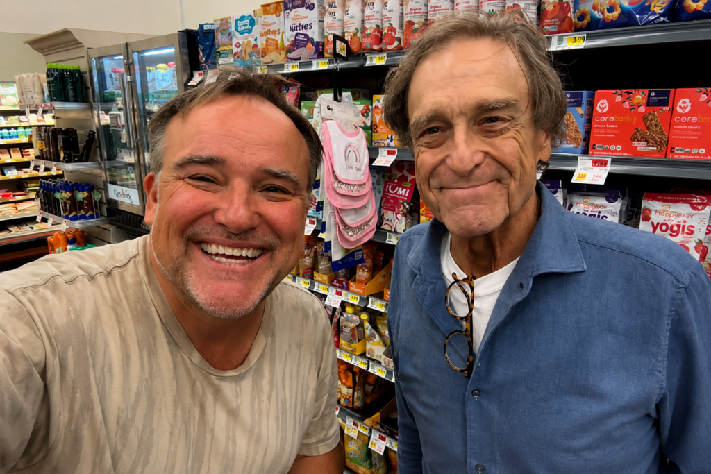 John Goodman smiles alongside actor David DeLuise in a supermarket aisle during a casual grocery store selfie.